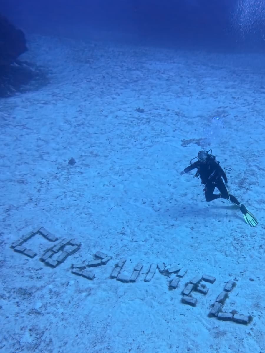 A scuba diver finning over a pale sand slope, above the word COZUMEL spelled out in stone blocks